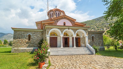 Grekisk ortodox kyrka nära Filippi, murad i grå sten med tre vita valv vid ingången och röda tegeltak. Bortanför finns en bergssluttning med buskvegetation.