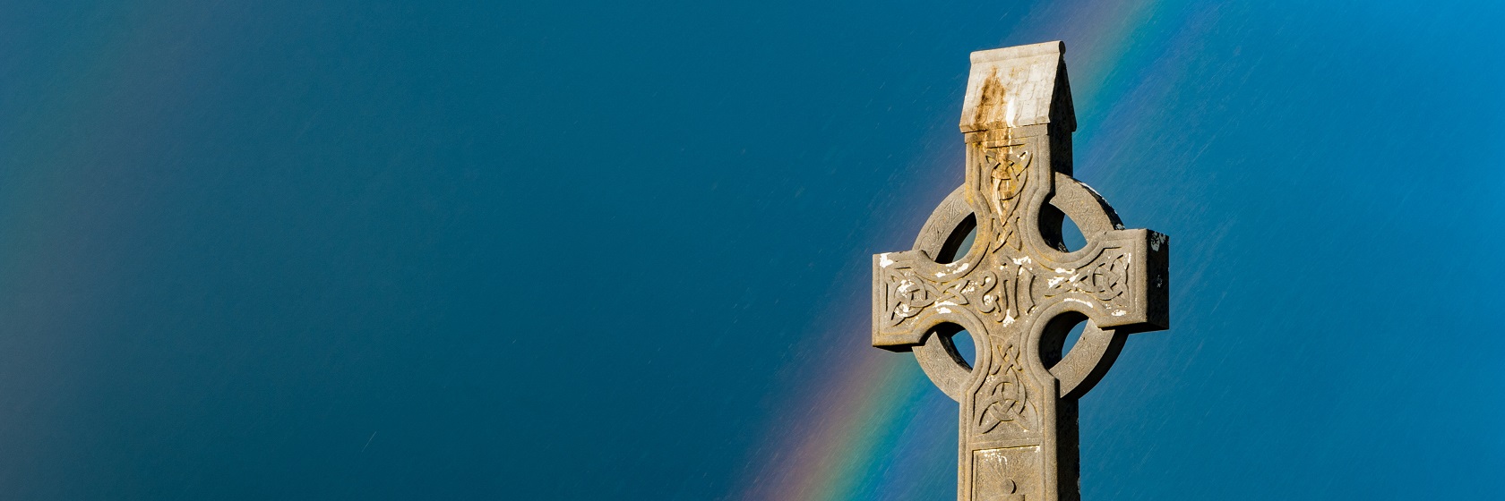 Ett ornamenterat keltiskt kors mot gråblå himmel, med en regnbåge som går genom korset.