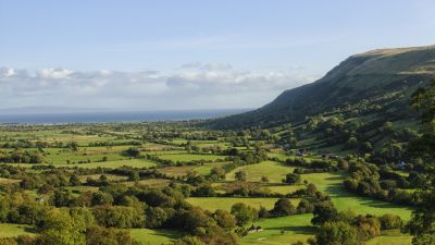 Ett vidsträckt irländskt landskap med ett lapptäcke av betesmarker avgränsade av trädrader samt en glest skogsklädd bergssluttning och havet i fjärran.