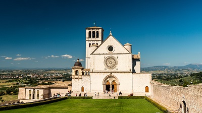 Franciscusbasilikan i Assisi lyser vitt i starkt solljus, kontrasterande mot en grn grsmatta och bl himmel.