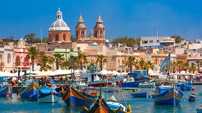 En malteisik hamn med färgglada småbåtar; i bakgrunden sticker tre rödaktiga kyrktorn upp ur bebyggelsen.