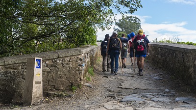 En grupp pilgrimsvandrare går bortåt över en bro, ledsagade av den gula pilgrimsmusslan på en låg stenpelare.