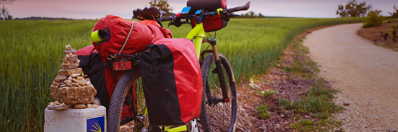 Mountain bike med röda cykelväskor parkerad vid en Caminovägsten, med ett grönt fält i bakgrunden.