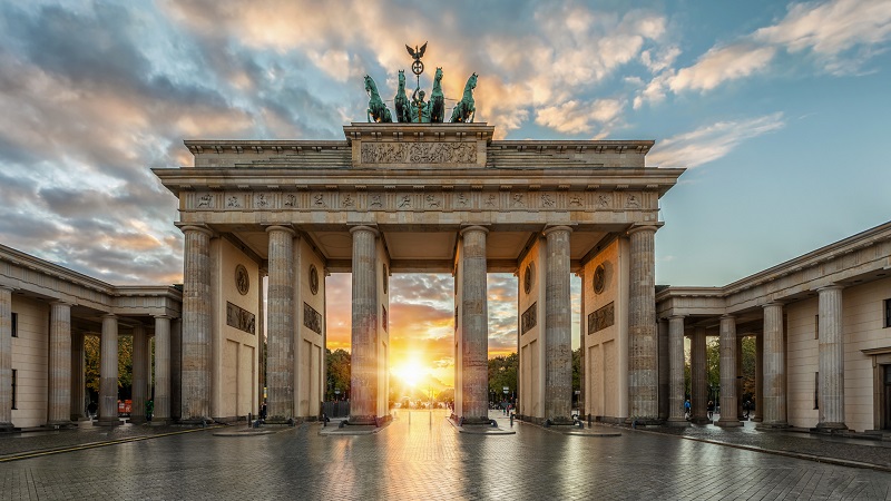 Solens sista strålar lyser genom Brandenburger Tor-monumentet i Berlin, mot en himmel med utdragna moln.
