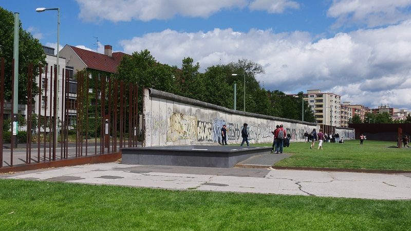 Del av Berlinmuren som målats med graffiti och människor som strövar omkring på gräset i närheten.