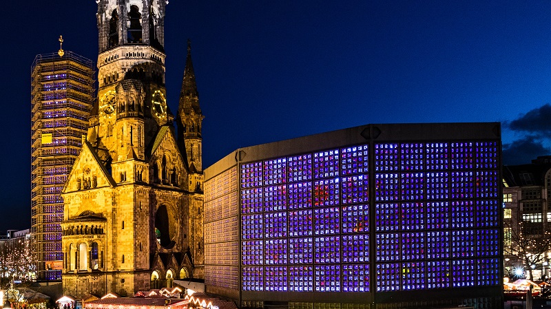 Resterna av Kaiser-Wilhelm-Gedächtniskirche i Berlin, som skadades svårt under de allierades bombningar av Berlin 1943. Intill finns en modern hexagonal kyrka vars många småfönster lyses upp i olika färger.